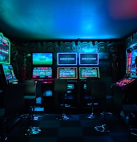 Dimly lit gaming arcade featuring a variety of brightly illuminated slot machines and video gaming screens. Neon signs and colorful displays create a vibrant atmosphere. The arrangement of gaming machines suggests an inviting space for entertainment, with chairs positioned for players.