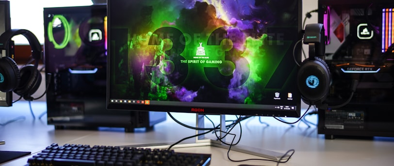 A gaming setup featuring a central monitor with a vibrant display showing 'The Spirit of Gaming'. On either side, there are gaming PCs with illuminated components and transparent cases. Headsets hang from the side of the monitor. The desk also has a black keyboard and mouse, and a branded mousepad.