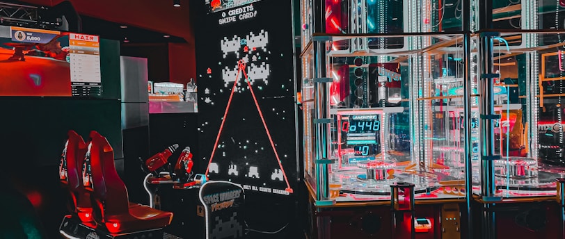 An arcade setting featuring a classic Space Invaders game setup and a modern gaming machine with seats. Bright neon lights illuminate the area, with scores displayed on digital screens. A bright and visually engaging environment full of electronic components.