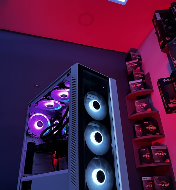 A gaming computer setup featuring a high-performance PC tower with multiple RGB fans lit in blue and purple hues. The case has a modern design with transparent panels showcasing internal components. In the background, shelves are stocked with various Ryzen and other gaming components under a vibrant red and blue ambient light.