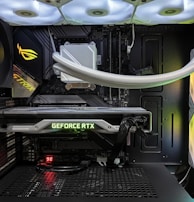 Inside view of a computer case showcasing a sophisticated gaming setup. Visible components include a GeForce RTX graphics card illuminated with green lighting, an ASUS motherboard, cooling fans with LED lighting, and a liquid cooling system with white tubes. Cables are neatly organized, and overall, the hardware suggests a high-performance machine.