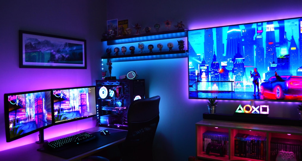 A vibrant gaming setup featuring multiple monitors displaying a cyberpunk cityscape. The room is illuminated with neon purple and blue LED lights, adding a futuristic ambiance. A custom-built PC with glowing components sits to the side, and shelves with collectible figurines are visible. A large digital screen on the wall shows an animated city scene, enhancing the immersive atmosphere.